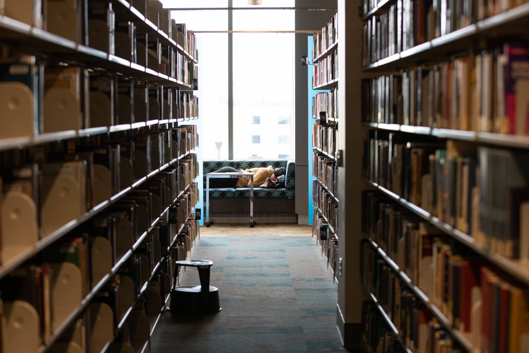 Person sleeping on library sofa at the end of hallway of bookshelves